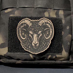 May include: A black and tan patch featuring a detailed illustration of a ram head with curved horns. The patch is centered on a black rectangular hook-and-loop fastener panel, which is attached to a dark camouflage patterned fabric surface.