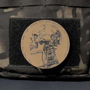 May include: A round, tan patch with a detailed illustration of a cyborg skull. The patch is affixed to a black square on a camouflage background. Text encircles the skull, reading "You may live to see man made horrors beyond your comprehension."