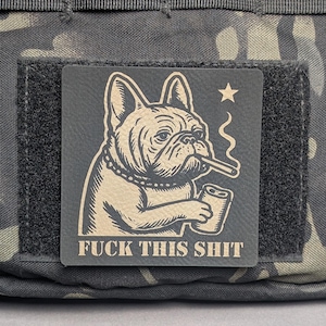 May include: A square patch featuring a bulldog smoking a cigarette and holding a can. The text "FUCK THIS SHIT" is printed below the bulldog. The patch is attached to a camouflage fabric.