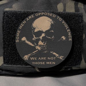 May include: A round patch with a skull and crossbones design. The patch is black with tan text that reads "SOME MEN ARE OPPOSED TO VIOLENCE" and "WE ARE NOT THOSE MEN". The patch is attached to a black fabric.