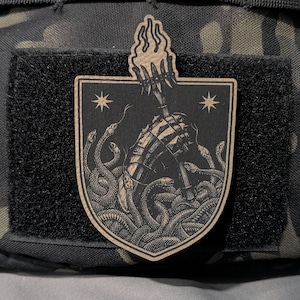 May include: A patch featuring a shield design with a skeletal hand holding a torch, surrounded by snakes. The patch is black with tan accents and two star symbols. The patch is attached to a black velcro surface.