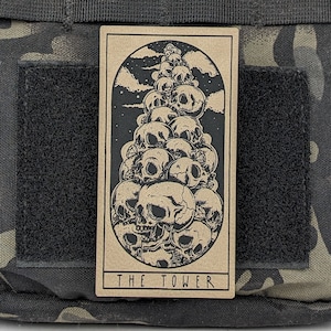 May include: A tarot card with a brown background and black ink illustration of a tower of skulls. The card reads "THE TOWER" at the bottom.