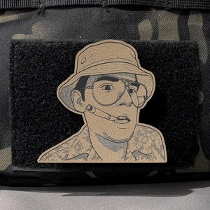 May include: A tan patch depicting a man in a bucket hat and aviator glasses, smoking a cigar. The patch is set on a black velcro base, with a camouflage pattern in the background.