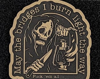 May include: A morale patch featuring a Grim Reaper holding a knife and a lantern. The text reads: "May the bridges I burn light the way" and "Fuck 'em all". The design is rendered in a tan and black color scheme with a die-cut border.