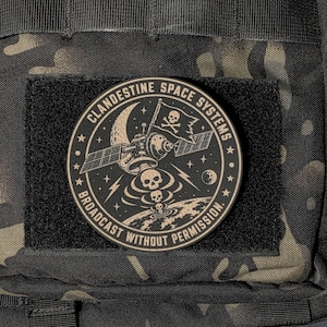 May include: A circular morale patch featuring a black and tan design with a satellite, a skull and crossbones flag, and a series of skulls descending toward Earth. The text reads CLANDESTINE SPACE SYSTEMS and BROADCAST WITHOUT PERMISSION.