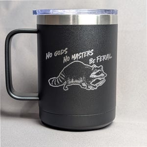 May include: Black stainless steel travel mug with a black handle. The mug has a white graphic of a raccoon holding a knife and the text "NO GODS NO MASTERS BE FERAL".