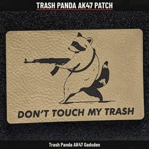 May include: A rectangular tan patch with a textured surface featuring a black graphic of a raccoon holding an AK-47 rifle. Below the raccoon, the text reads "DON'T TOUCH MY TRASH" in bold black capital letters. The design is centered.