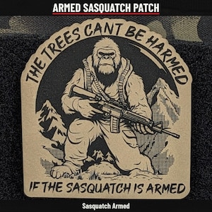 May include: A tan and black morale patch featuring a graphic of a Sasquatch holding a rifle in a mountainous landscape. The text reads THE TREES CANT BE HARMED IF THE SASQUATCH IS ARMED. The design is circular with a scalloped border edge.