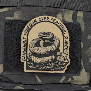 May include: A tan-colored patch with a black snake coiled around a flower. The patch has the text "Dangerous Freedom Over Peaceful Slavery" around the snake.