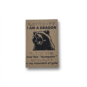May include: A rectangular, tan-colored sticker with Japanese text and the English phrase "I AM A DRAGON." Below, a black and white illustration of a raccoon with its mouth open. The text "and this 'dumpster' is my mountain of gold" is also present.