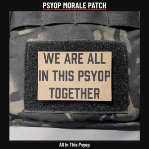 May include: A rectangular morale patch with a tan background and black text that reads WE ARE ALL IN THIS PSYOP TOGETHER. The patch is attached to a black hook-and-loop fastener strip on a dark camouflage fabric surface.