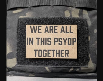 Könnte beinhalten: Ein rechteckiger Aufnäher mit beigem Hintergrund und schwarzem Text, der WE ARE ALL IN THIS PSYOP TOGETHER lautet. Der Aufnäher ist auf einem schwarzen Klettstreifen auf einem dunklen Tarnstoff befestigt.
