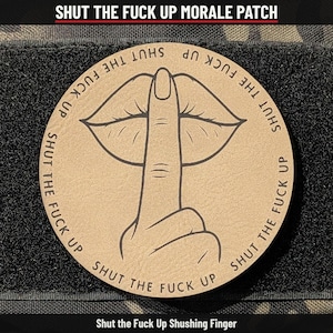 May include: A circular tan morale patch featuring a dark brown line art illustration of lips with a finger pressed against them in a shushing gesture. The text SHUT THE FUCK UP is repeated in a circular pattern around the central graphic.