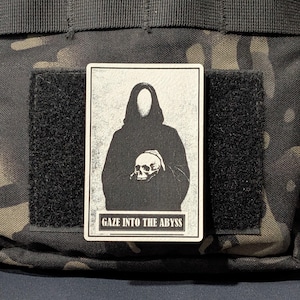 May include: A rectangular patch with a black and white illustration of a hooded figure holding a skull. The text "GAZE INTO THE ABYSS" is printed below the image. The patch is attached to a black velcro square.