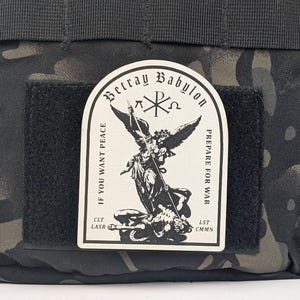 May include: A black and white patch with an angel holding a spear and a demon lying beneath it. The patch has the text "Betray Babylon" at the top, "If you want peace" on the left, and "Prepare for war" on the right. The patch also has the text "CLT LASR" on the bottom left and "LST CMNN" on the bottom right.