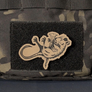 May include: A tan-colored morale patch featuring a black graphic illustration of a dead rat with an X over its eye, positioned on a black rectangular hook-and-loop fastener base, which is attached to a dark camouflage patterned fabric surface.