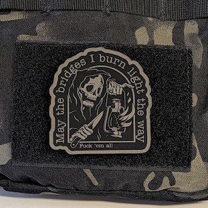 May include: A black patch with a Grim Reaper design holding a lantern and knife. The text reads "May the bridges I burn light the way" and "Fuck 'em all". The patch is attached to a camouflage fabric.