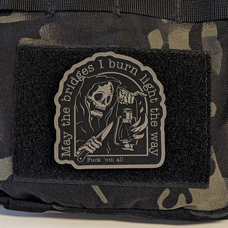 May include: A black patch with a Grim Reaper design holding a lantern and knife. The text reads "May the bridges I burn light the way" and "Fuck 'em all". The patch is attached to a camouflage fabric.