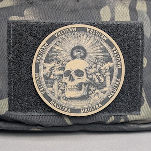 May include: A round patch with the word "MKULTRA" encircling a central design. The design features a skull, an eye, and radiating lines. The patch is attached to a black velcro square, set against a camouflage background.