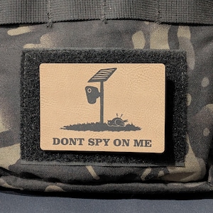May include: A rectangular tan morale patch with a black hook-and-loop backing attached to a camouflage fabric. The patch features a black graphic of a solar-powered security camera and a dead bird, with the text DONT SPY ON ME printed below.