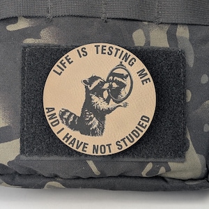 May include: A brown leather patch with the text "Life is testing me and I have not studied" and a black and white illustration of a raccoon looking in a mirror.