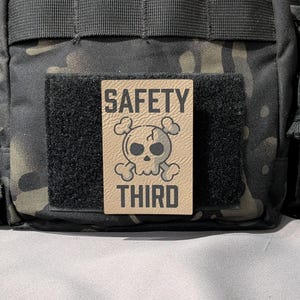 May include: Tan rectangular patch with the words "SAFETY THIRD" and a skull and crossbones graphic. The patch is attached to a black velcro square, which is on a camouflage background.