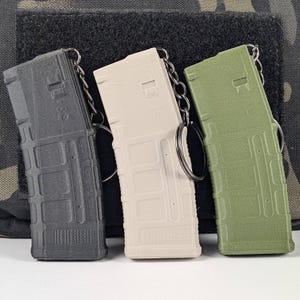 May include: Three plastic magazine keychains in black, beige, and green. The keychains are shaped like rifle magazines and have a silver key ring.