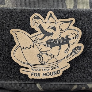 May include: Tan and black patch featuring a cartoon fox holding a gun and grenade, with a bullet in flight. The text reads "Special Force Group FOX HOUND". The patch is on a black and camouflage background.