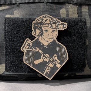 May include: A tan and black patch depicting a stylized illustration of a person wearing a helmet with night vision goggles, a headset, and holding a rifle. The patch is attached to a black velcro surface, against a camouflage background.