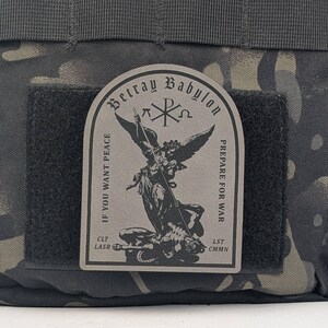 May include: A black and gray tactical bag with a patch that reads "Betray Babylon" and features an angel with a spear fighting a demon. The patch also includes the text "If you want peace prepare for war" and "CLT LASR LST CMN".