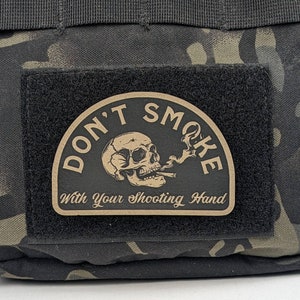 May include: A brown patch with black stitching and the text "Don't Smoke With Your Shooting Hand" in a white font. A skull with a cigarette in its mouth is depicted inside a black circle.