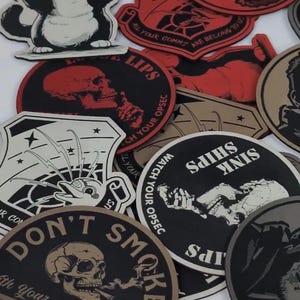May include: A collection of black and white, red, and tan round patches with various designs. The patches feature skulls, a cat, and text that reads "Don't Smoke", "Ships Sink", "Watch Your Opsec", and "All Your Comms Are Belong To Us".