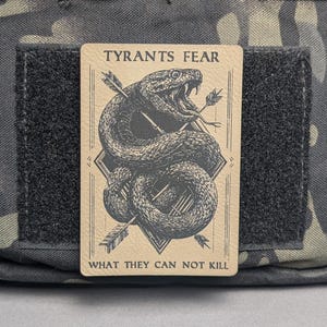 May include: A rectangular patch featuring the text "TYRANTS FEAR WHAT THEY CAN NOT KILL" and a detailed illustration of a coiled snake with arrows. The patch is tan with black text and a black and grey snake design. The patch is attached to a camouflage background.