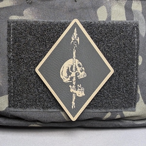May include: A diamond-shaped patch featuring a skull pierced by a spear. The skull and spear are tan, set against a black background. The patch is attached to a black velcro square, which is on a camouflage fabric.