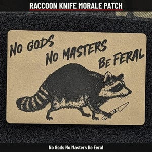 May include: A rectangular morale patch with a tan background and black graphic print. The design features a raccoon holding a small knife, accompanied by the text "NO GODS NO MASTERS BE FERAL" printed in a bold, black, sans-serif font above the animal.