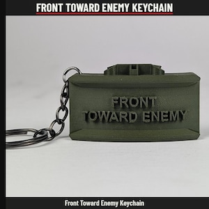 May include: An olive green rectangular keychain featuring the raised text FRONT TOWARD ENEMY on the front surface. The keychain is attached to a dark metal chain and a circular split ring, mimicking the design of a military mine.