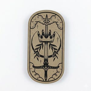 May include: A tan, oval sticker featuring a black illustration of a sword, crown, and olive branches. The design is framed by clouds and stars. The sword is central, with the crown above and branches on either side.