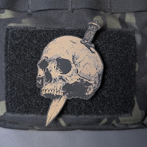May include: A tan and black patch featuring a skull pierced by a knife. The skull is detailed with shading and cracks, and the knife has a textured handle. The patch is set against a black square on a camouflage background.