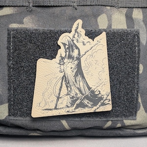 May include: A tan patch featuring a Grim Reaper illustration with a lantern and staff, set against a smoky background. The patch is affixed to a black velcro square on a camouflage fabric.