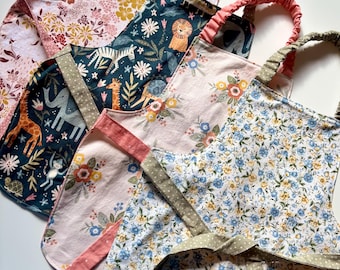 May include: A collection of four aprons featuring various patterns including a dark blue fabric with jungle animals, a light pink fabric with floral designs, and a white fabric with small blue and yellow flowers, all with elasticized neck straps.
