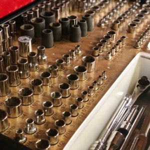 Può includere: Una cassetta degli attrezzi in legno con una varietà di chiavi a bussola in metallo organizzate per dimensione. Le bussole sono disposte in file e colonne, con ogni fila etichettata con una frazione. La cassetta degli attrezzi ha anche un cassetto in plastica bianca con un set di cacciaviti.