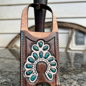 May include: A brown leather knife sheath with a turquoise and silver decorative design. The sheath has a handle loop and is stitched with teal thread. The knife handle is dark brown.