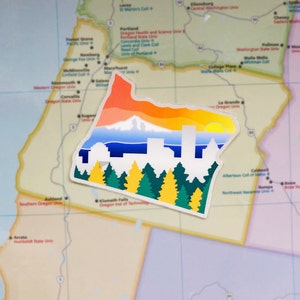 Puede incluir: Una pegatina del estado de Oregón con una puesta de sol colorida, una cordillera y una ciudad. La pegatina está colocada en un mapa de los Estados Unidos.