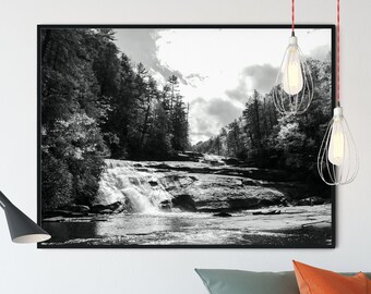 Könnte beinhalten: Schwarzweißfoto eines Wasserfalls, der über Felsen in einer Waldlandschaft herunterstürzt. Das Bild ist in einem schwarzen Rahmen gerahmt und hängt an einer weißen Wand über einem grünen Sofa mit orangefarbenen Kissen.