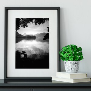 May include: A black and white photograph of a lake with mountains in the background. The photo is framed in a black frame and is hanging on a white wall. A small green plant in a white pot with a leaf pattern is sitting on a black shelf below the photo.