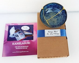 May include: A Kintsugi repair kit featuring a blue ceramic bowl with gold-colored cracks, a brown cardboard box with a blue ribbon and a label reading Kintsugi Planet The Art of Kintsugi, and a pink instruction manual labeled KANELASURI.