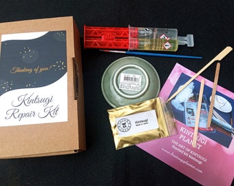 May include: A Kintsugi repair kit featuring a brown cardboard box with a label reading Thinking of you! Kintsugi Repair Kit, a red dual-syringe applicator, a blue paintbrush, a small green ceramic dish, a gold powder packet, and wooden sticks.