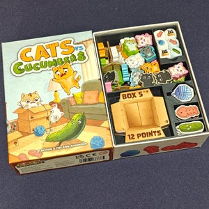 May include: Opened board game box titled "Cats vs Cucumbers." The box art features cartoon cats, a cucumber, and the game's title. Inside, the game includes cat-shaped game pieces, cucumber tokens, and a box labeled "BOX 5" with "12 POINTS."
