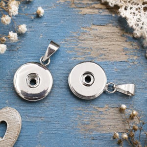 Könnte beinhalten: Zwei runde, silberfarbene Druckknopf-Anhänger aus Metall. Jeder Anhänger hat eine polierte, reflektierende Oberfläche mit einem zentralen, kreisförmigen Druckknopfmechanismus und einer kleinen, dreieckigen Öse an der Oberseite.