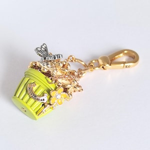 May include: A Juicy Couture charm in the shape of a green bucket with gold and yellow floral accents. The charm has a gold-colored clasp and chain. The text "Juicy Couture" is visible on the bucket.